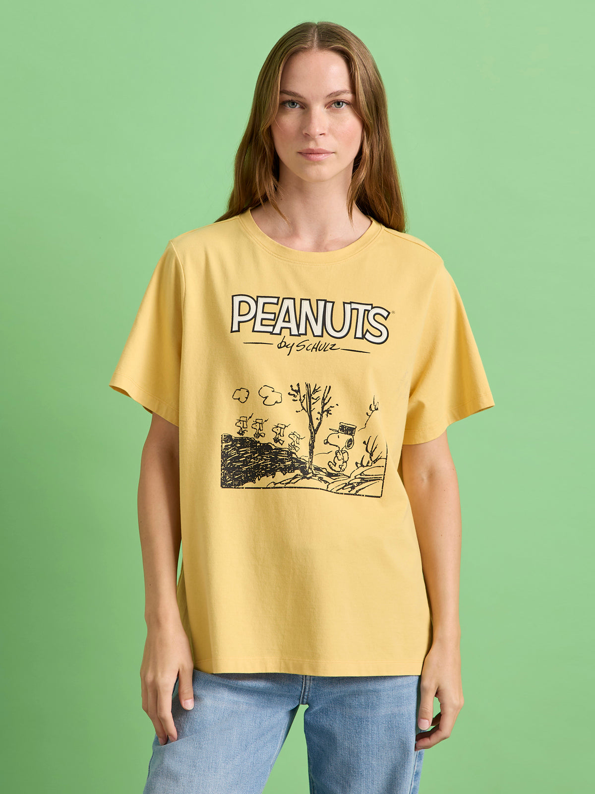 A person with long brown hair wears the Sanctuary Clothing x PEANUTS 75th Anniversary marching tee in washed gold, featuring a black and white Snoopy comic illustration, standing against a green background.