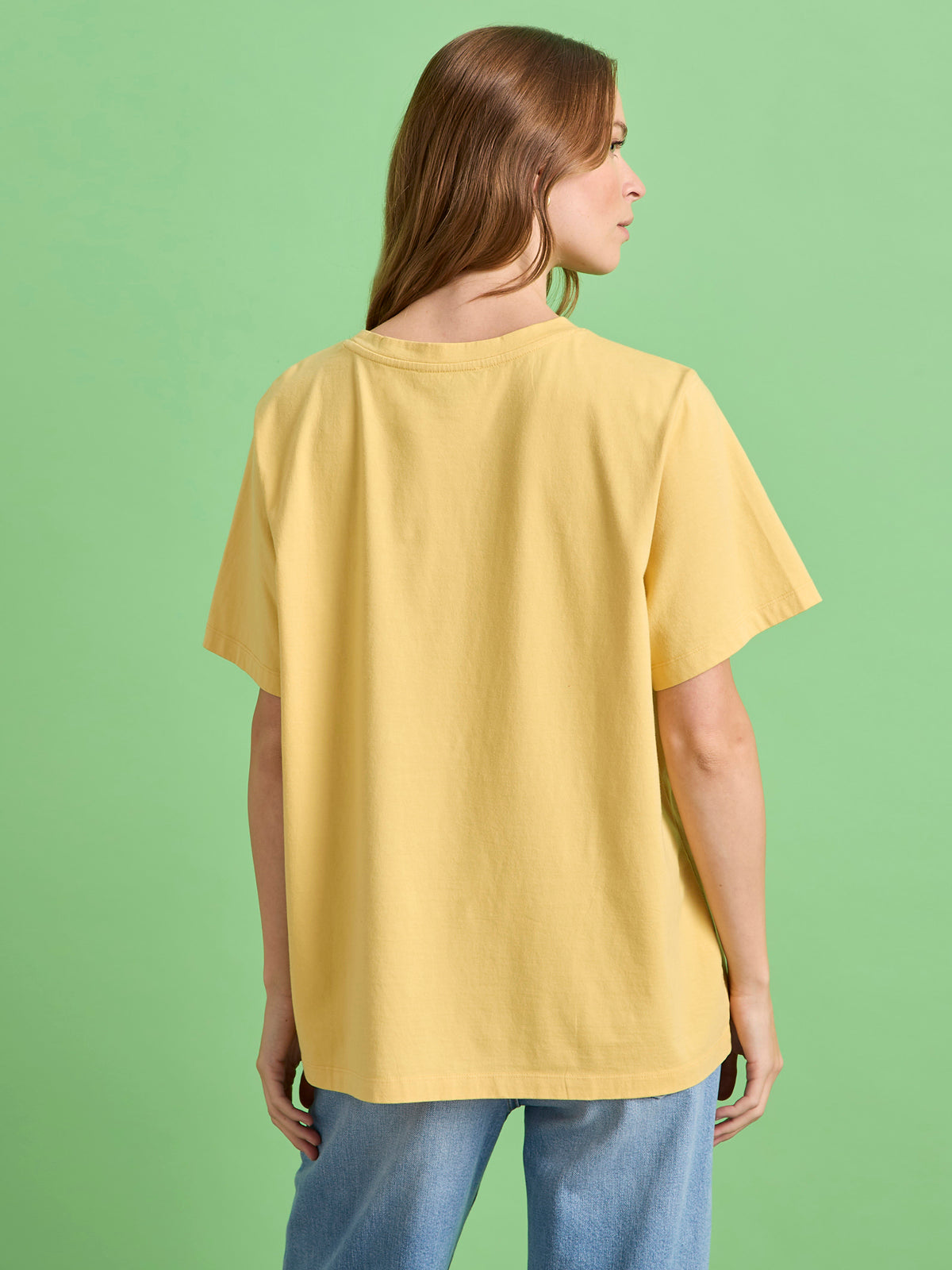 A woman with light brown hair faces away from the camera, wearing the Sanctuary Clothing x PEANUTS 75th Anniversary marching tee in washed gold and blue jeans, standing against a plain green background.