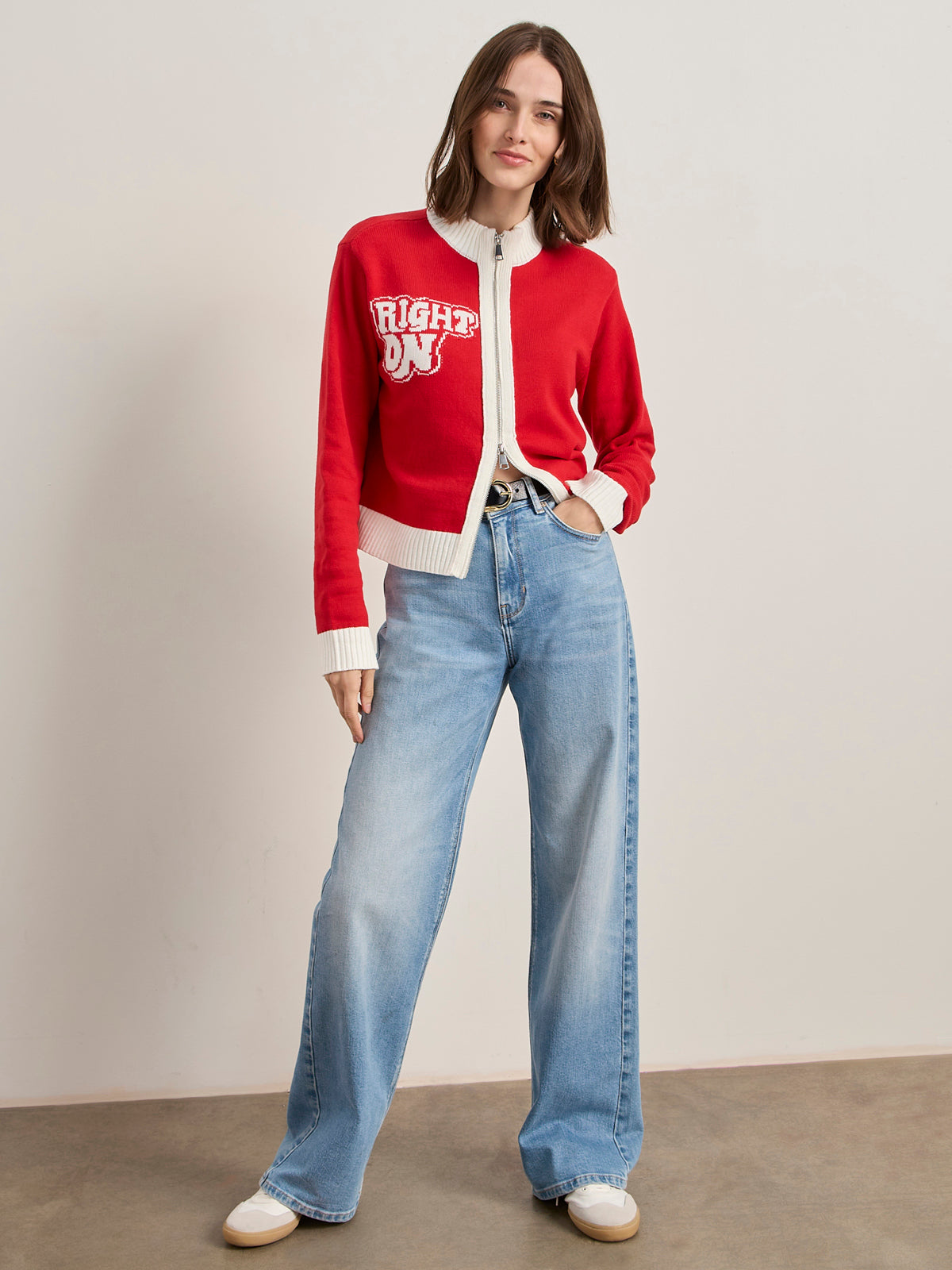 A woman poses against a plain wall, wearing the joe cool zip up sweater cherry lacquer by Sanctuary Clothing x PEANUTS 75th Anniversary with light blue high-waisted wide-leg jeans and white sneakers, smiling softly at the camera.