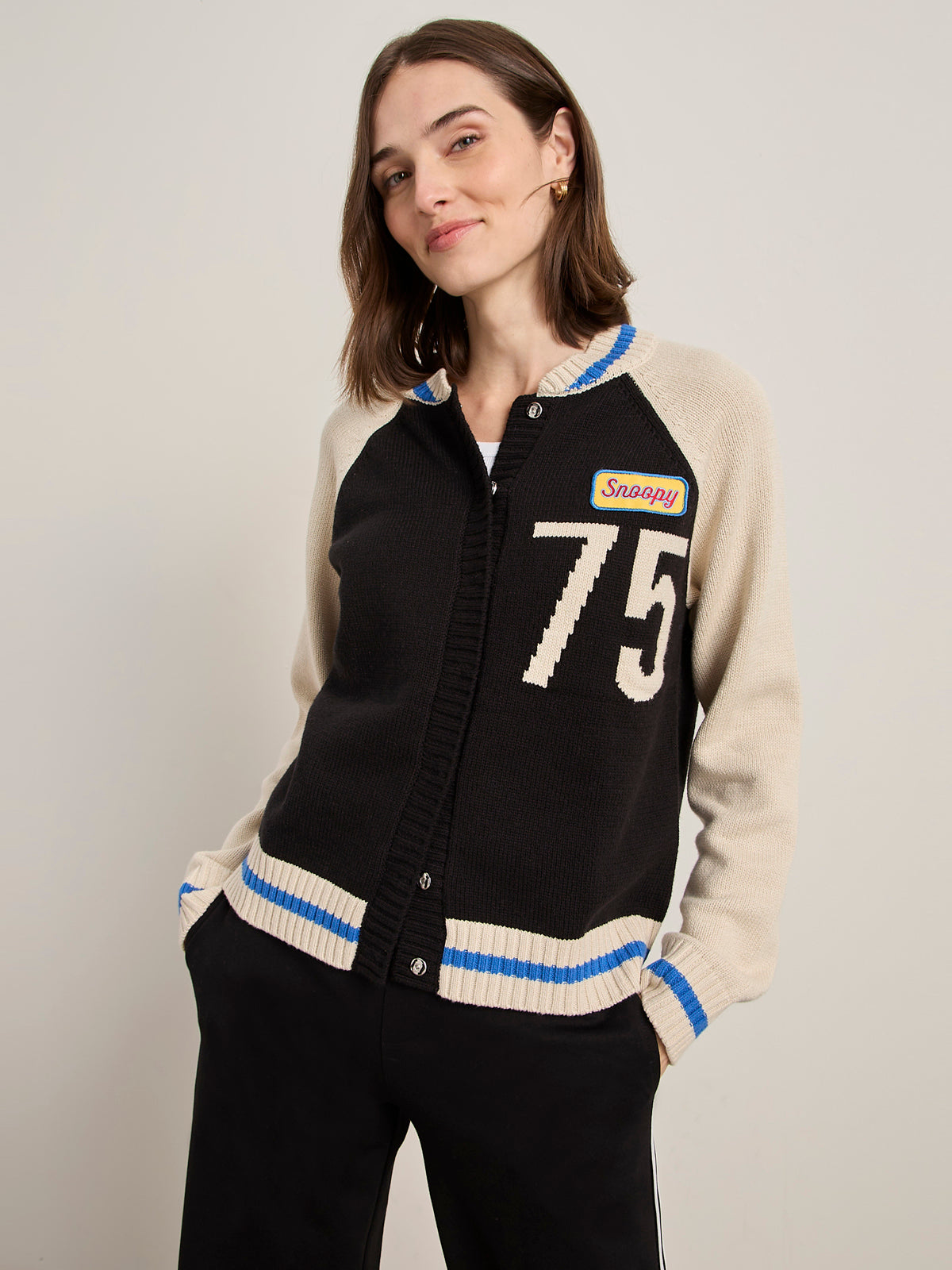 A woman with shoulder-length brown hair wears the Sanctuary Clothing x PEANUTS 75th Anniversary "75 bomber sweater black chalk," featuring blue stripes, a "Snappy" patch, and the number 75. She smiles slightly against a plain background.