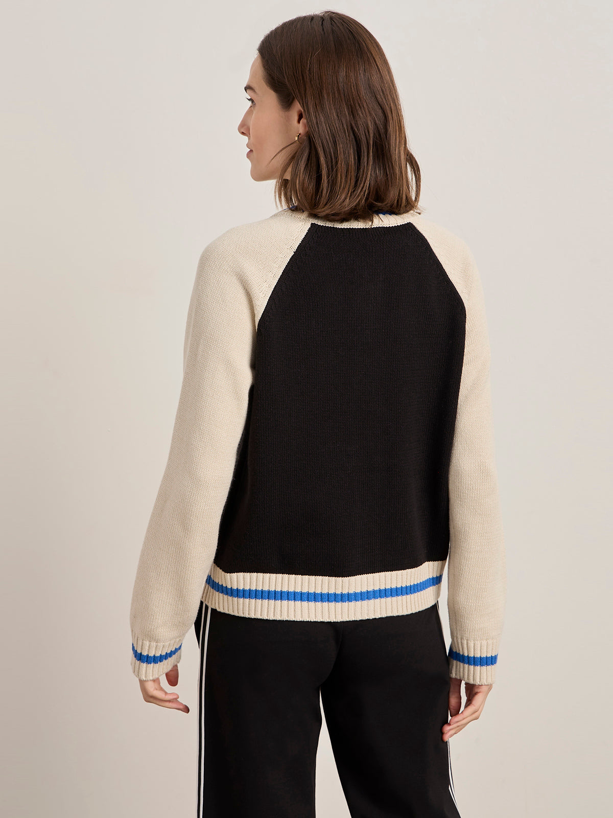 A person with shoulder-length brown hair faces away from the camera, wearing the Sanctuary Clothing x PEANUTS 75th Anniversary 75 bomber sweater black chalk and black pants with white side stripes.