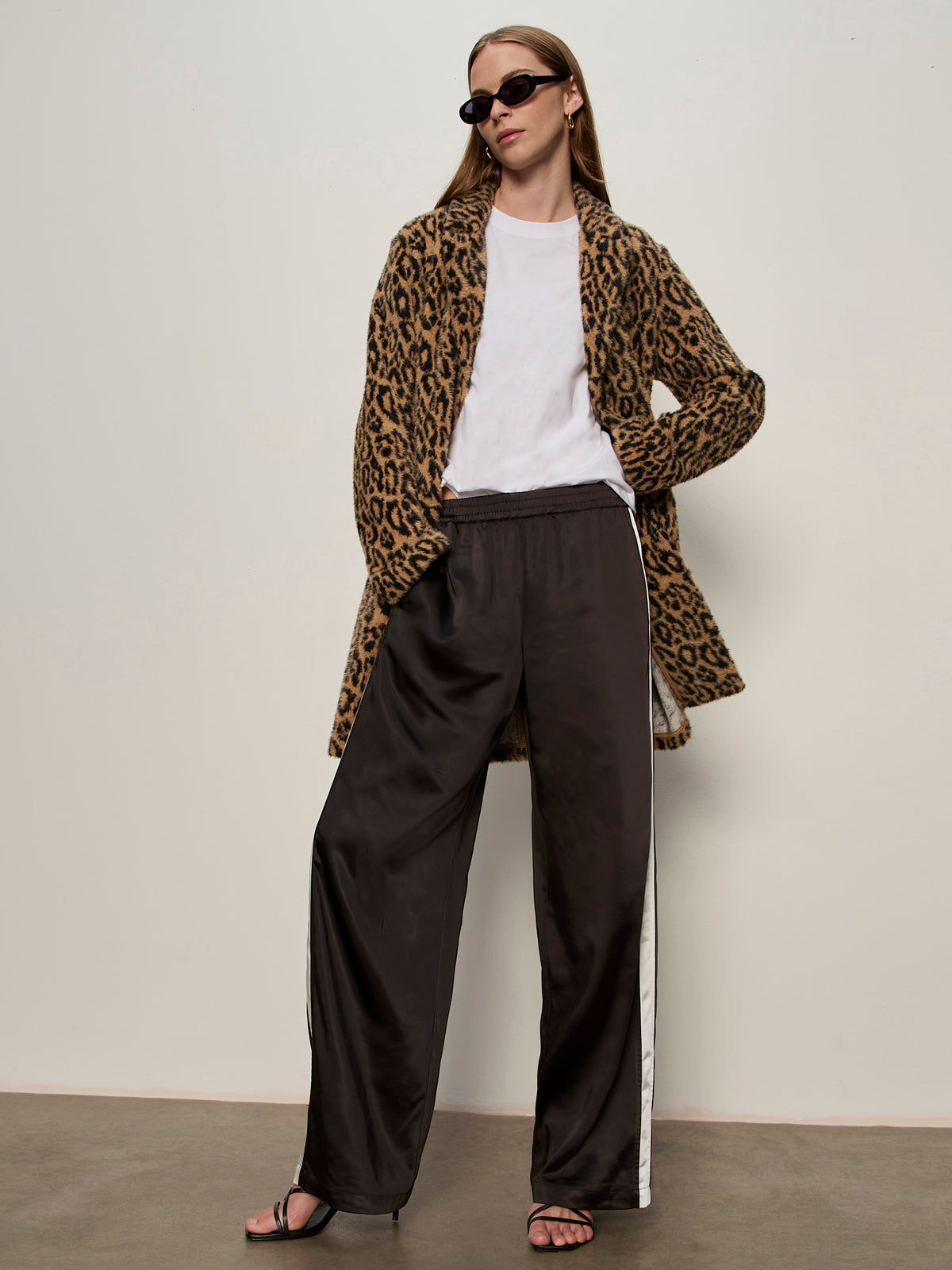 A person stands confidently against a plain background wearing black sunglasses, a white t-shirt, Sanctuary Clothing„¢s hometown jacket lynx, loose black pants with white side stripes, and black heeled sandals.