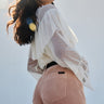 A woman with wavy hair stands outdoors in sunlight, wearing a sheer white blouse with ruffled sleeves and Sanctuary Clothing's corduroy marine pant in dusty rose.