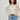 A woman with curly hair wears the Sanctuary Clothing boho lace top vintage ivory over a white top, tucked into light blue jeans with a brown belt, standing against a plain light-colored wall.