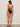 A woman with long brown hair, wearing black sandals, faces away from the camera in a Sanctuary Clothing opened front mio one piece swimsuit in black, featuring thin shoulder straps and crisscross lace-up back detailing.