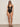A woman models the Sanctuary Clothing opened front mio one piece swimsuit in black, featuring two front cutouts with decorative buckles. She pairs it with black sandals and sunglasses against a plain light background.