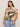 A woman in Sanctuary Clothing's "the perfect tee green vista extended sizing"—a short-sleeve top with an abstract pink, green, and beige leaf pattern—stands against a plain background, wearing light blue jeans and gold hoop earrings.
